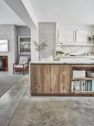 Et hvidt, rustikt køkkendesign med poleret betongulv, en køkkenø designet i træ med hvide marmorbordplader og lysegrå skabe.
