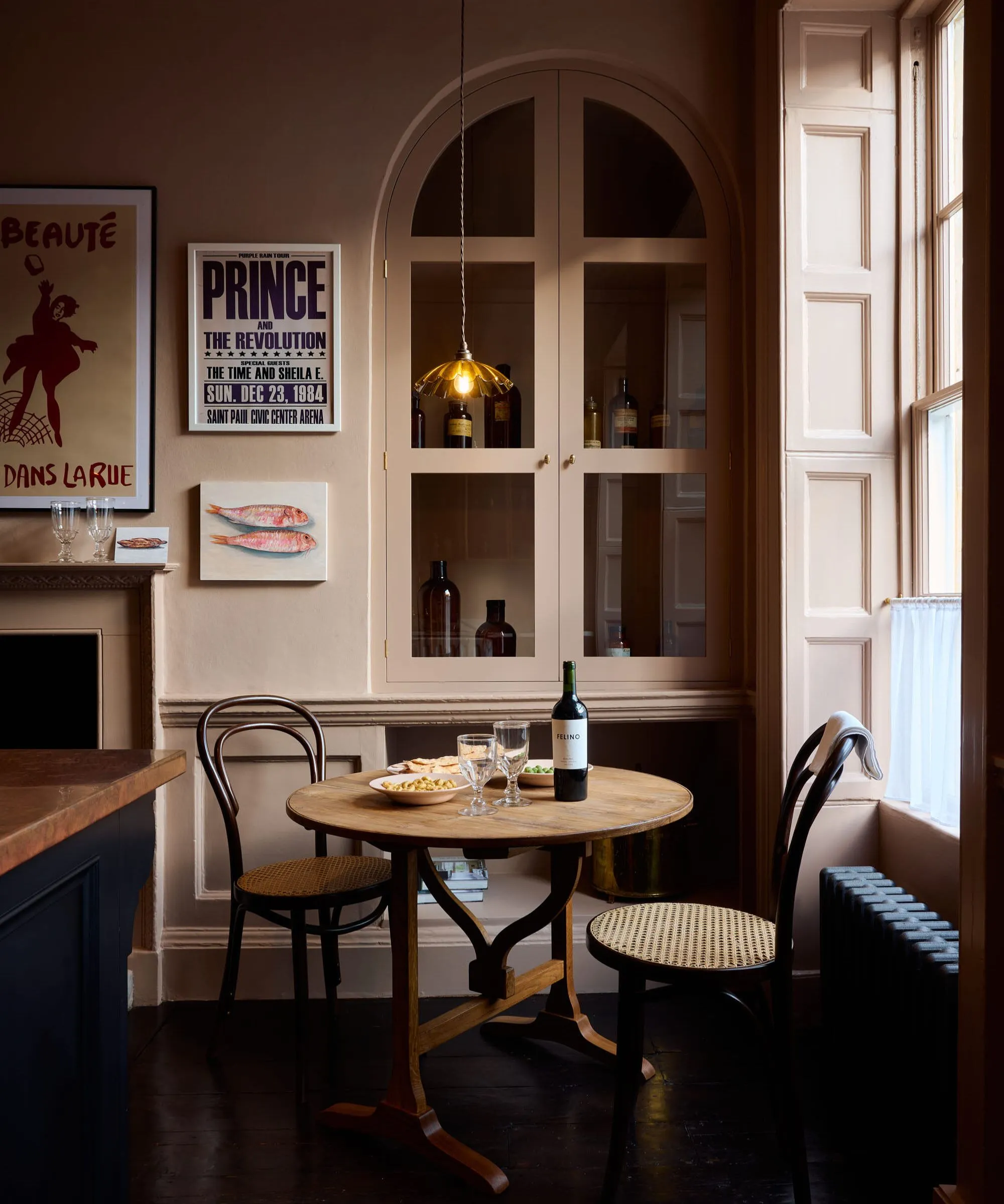 Kuchnia w stylu francuskiego bistro z małym okrągłym stołem, drewnianymi krzesłami bistro, różową ścianą, szafką i niską lampą wiszącą' loading='lazy' title=