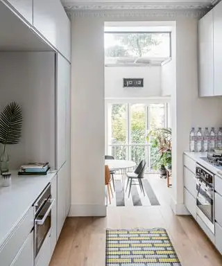 Una cocina de galera blanca con puertas sin tiradores que dan a un comedor con grandes ventanales.