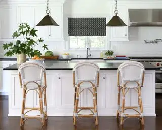Cocina blanca y luminosa con una gran isla con encimera de granito negro y base blanca con alacenas, tres sillas de bar de ratán y dos luces colgantes de cono negro colgando sobre la isla.