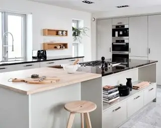 Cocina moderna con gran isla, encimera de madera clara y piedra natural antigua negra, armarios de color gris pálido y paredes pintadas de blanco, madera clara y taburete redondeado.