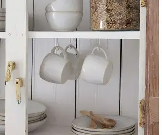 Tazas de cerámica blanca colgadas de ganchos debajo de un estante de gabinete con platos blancos anidados y cucharas de madera a la vista