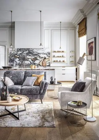 zona giorno della cucina con pareti e mobili bianchi, dettagli dorati, piano di lavoro e alzatina in marmo, lampade a sospensione, divano grigio, tappeto oro e bianco sporco, tavolini metallici, pavimento in legno duro