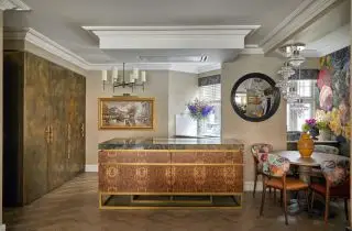 Uma cozinha, área de jantar e de estar com armários marrons do chão ao teto, uma ilha vermelha e dourada e uma mesa circular com cadeiras florais