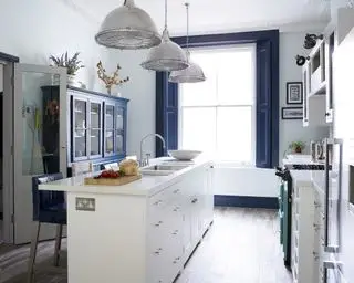 Uma estreita ilha de cozinha branca em frente a uma grande janela azul e luzes pendentes de metal