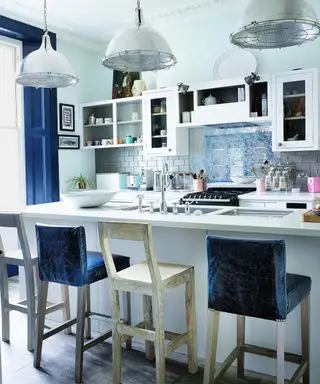 Um balcão de café da manhã branco com bancos azuis e brancos abaixo de três luminárias pendentes de metal industrial