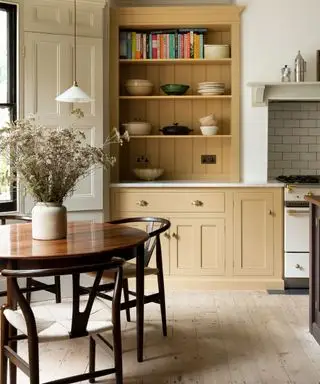 Cocina estilo shaker de color amarillo con una mesa redonda de madera y sillas con un gran jarrón de flores en el centro