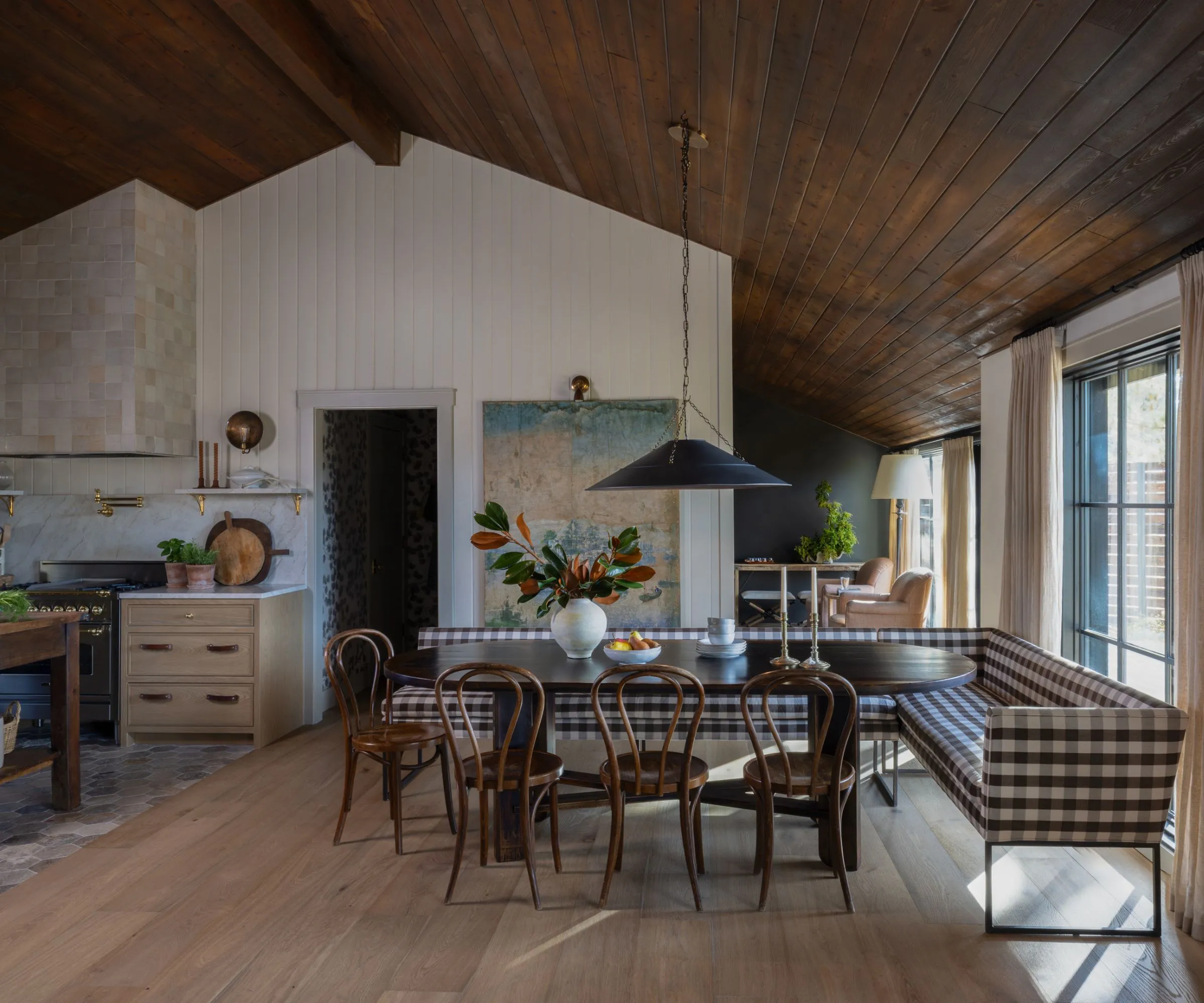 Eine Essecke mit einem dunklen ovalen Holztisch, umgeben von Holzstühlen und einer karierten Sitzbank. An einer Wand hängt ein großer Vintage-Wandbehang und über dem Tisch hängt eine schwarze Pendelleuchte' loading='lazy' title=