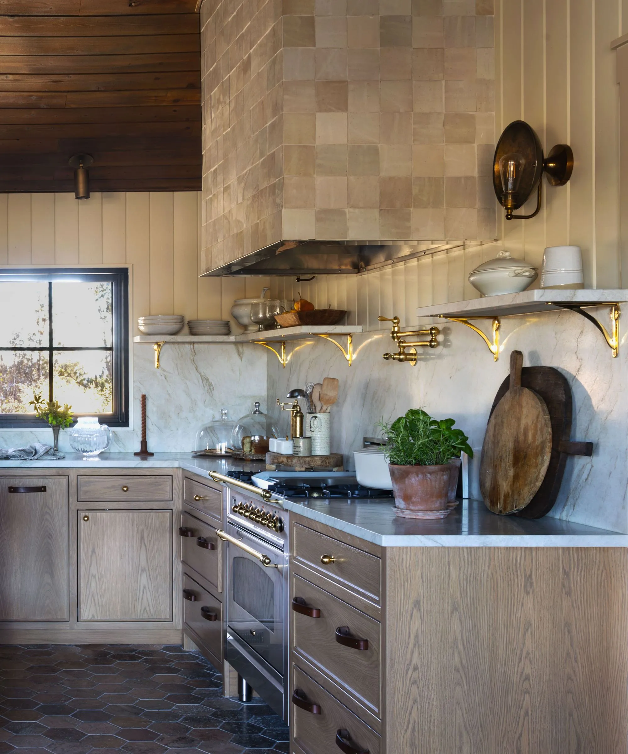 Eine sonnendurchflutete Frühstücksecke mit einer geschwungenen cremefarbenen Einbaubank und einem runden Holztisch. Darüber hängt eine Messingpendelleuchte, eingerahmt von einem großen Bogenfenster und schwarzen Glastüren mit Stahlrahmen' loading='lazy' title=