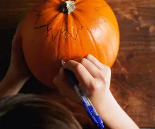 Zwei Hände zeichnen einen Umriss auf einem großen orangefarbenen Kürbis