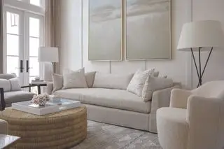 sala de estar color hueso con una gran mesa de centro redonda de ratán, bandeja blanca, obras de arte, lámparas de pie blancas, lámpara de mesa y sillón boucle