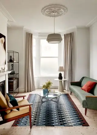 sala de estar blanca con muebles modernos de mediados de siglo, alfombra gráfica, mesa de café de vidrio, sofá verde, sillón amarillo, colgante, obras de arte y almacenamiento retro