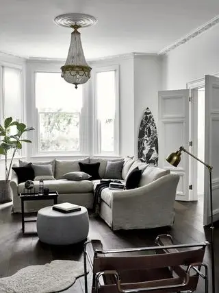 sala de estar branca com sofá secional cinza, piso de madeira escura, pufe, detalhes em preto, janela saliente