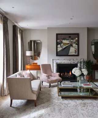 Ein stilvolles Wohnzimmer in mittlerem Grau-Beige, mit Spiegeln und Strahlern und einem Couchtisch aus Glas vor einem Kamin.