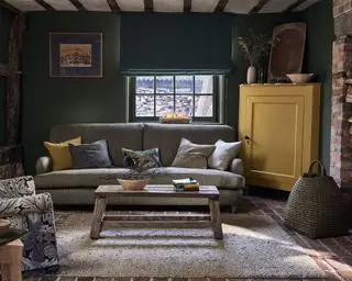 Dunkelgrün gestrichenes Wohnzimmer mit gelb gestrichenen Möbeln, grauem Sofa, Loungesessel und Kissen mit William Morris-Print, dunklen Holzelementen