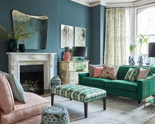 Wohnzimmer mit grünem Sofa und verschiedenen Kissen sowie gepolstertem Couchtisch mit Blattmuster und Schiebefenster mit hellen Kissen und blaugrünen Wänden