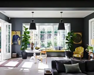Ein dunkelgrau gestrichenes Wohnzimmer mit senffarbenen Akzenten und großen hellen Fenstern.