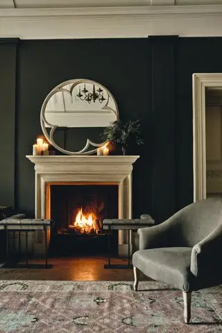 Schwarzes Wohnzimmer mit großem runden Spiegel, offenem Kamin, Vintage-Teppich, grauem Sessel, brennenden Kerzen auf dem Kaminsims und Vase mit Blattwerk