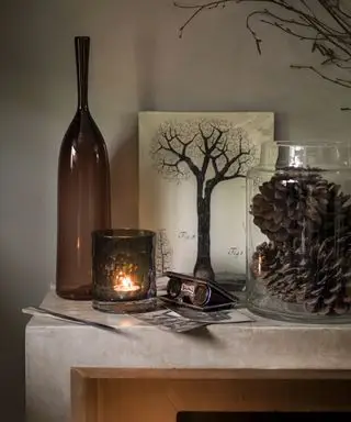Herbstliche Kaminsims-Ideen mit einer braunen Glasflasche und einem Kerzenhalter, Tannenzapfen in einer Hurrikan-Vase und einem Baumgemälde
