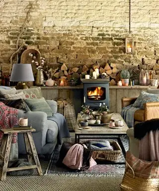 Herbstliche Kaminsims-Ideen mit einer freiliegenden Steinwand, einem Ofen, grauen Sofas mit Decken und Brennholz und unpassenden Ornamenten auf dem Kaminsims