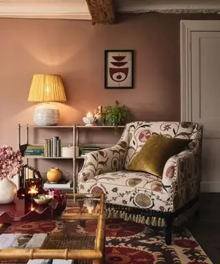 Landhaus mit rosa gestrichenen Wänden, Holzbalken an der Decke, Konsolentisch aus Metall mit Tischlampe, dekoriert mit Büchern und Ornamenten, gepolsterter Sessel mit Blumenmuster und Quasten, Teppich mit Blumenmuster auf dunklem Holzboden, Couchtisch aus Bambus, Glas und Rattan, dekoriert mit rotes Tablett mit Kerze, Vase mit Blumen und Schmuckstücken