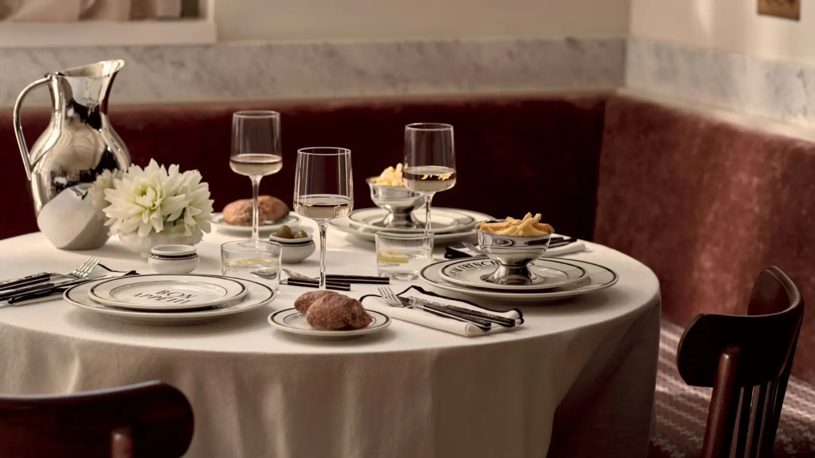 Una mesa de bistró con un mantel blanco, platos con borde negro, tres copas de vino y una jarra de acero inoxidable.' loading='lazy' title=