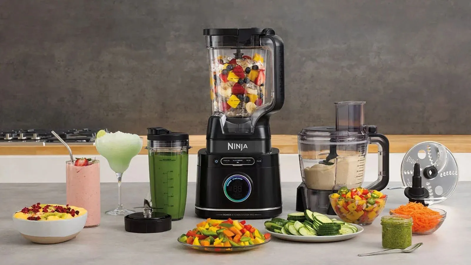 En Ninja blender fylt med frukt; en Ninja food prosessor som lager en dip; omgitt av tallerkener med kuttede grønnsaker og glass med blandet frukt, alt på en hvit disk mot en grå bakgrunn.' loading='lazy' title=