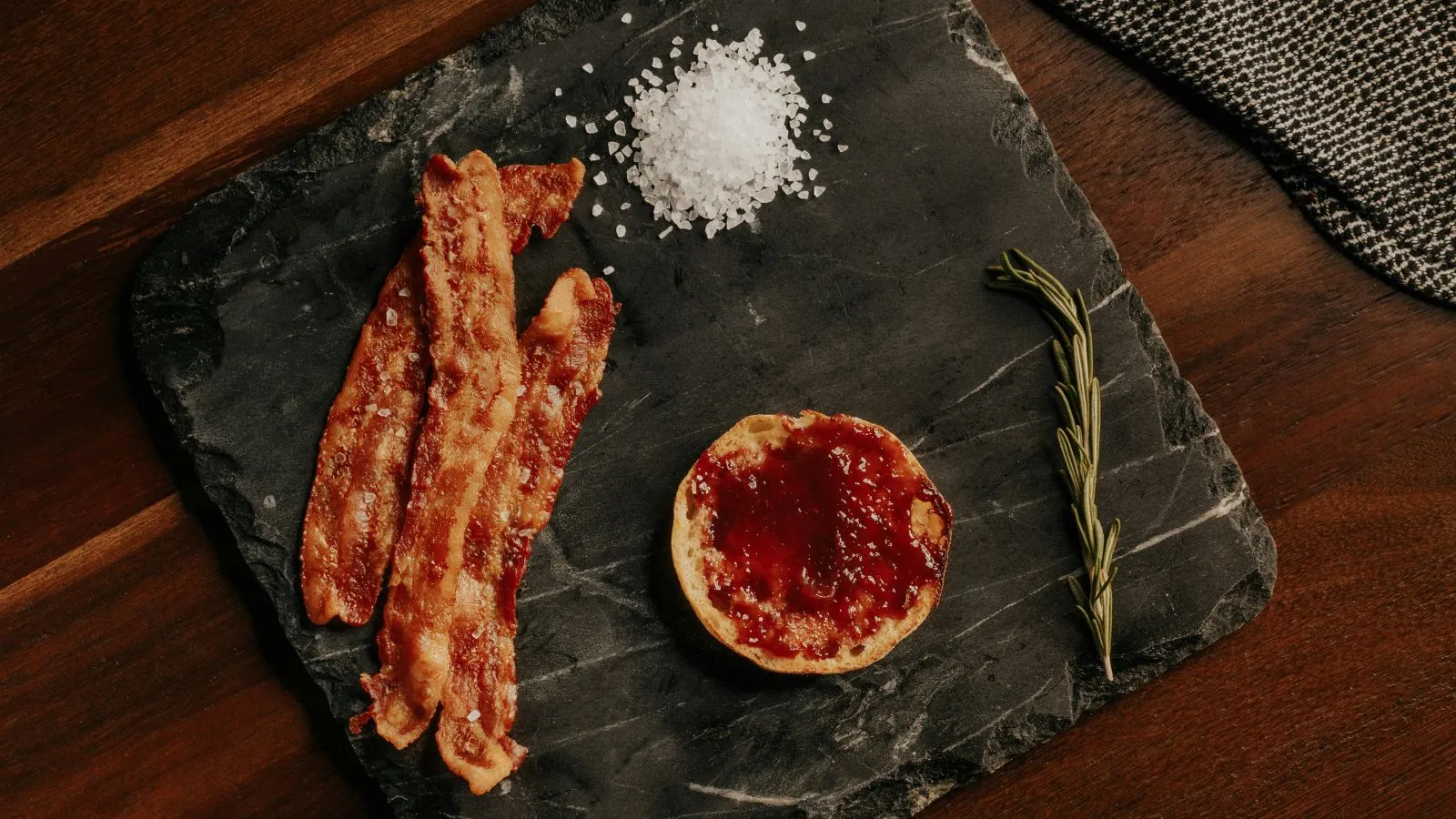 Tres erupciones de tocino en una bandeja de pizarra acompañada de un manantial de romero una pequeña porción de sal y una olla de salsa roja' loading='lazy' title=