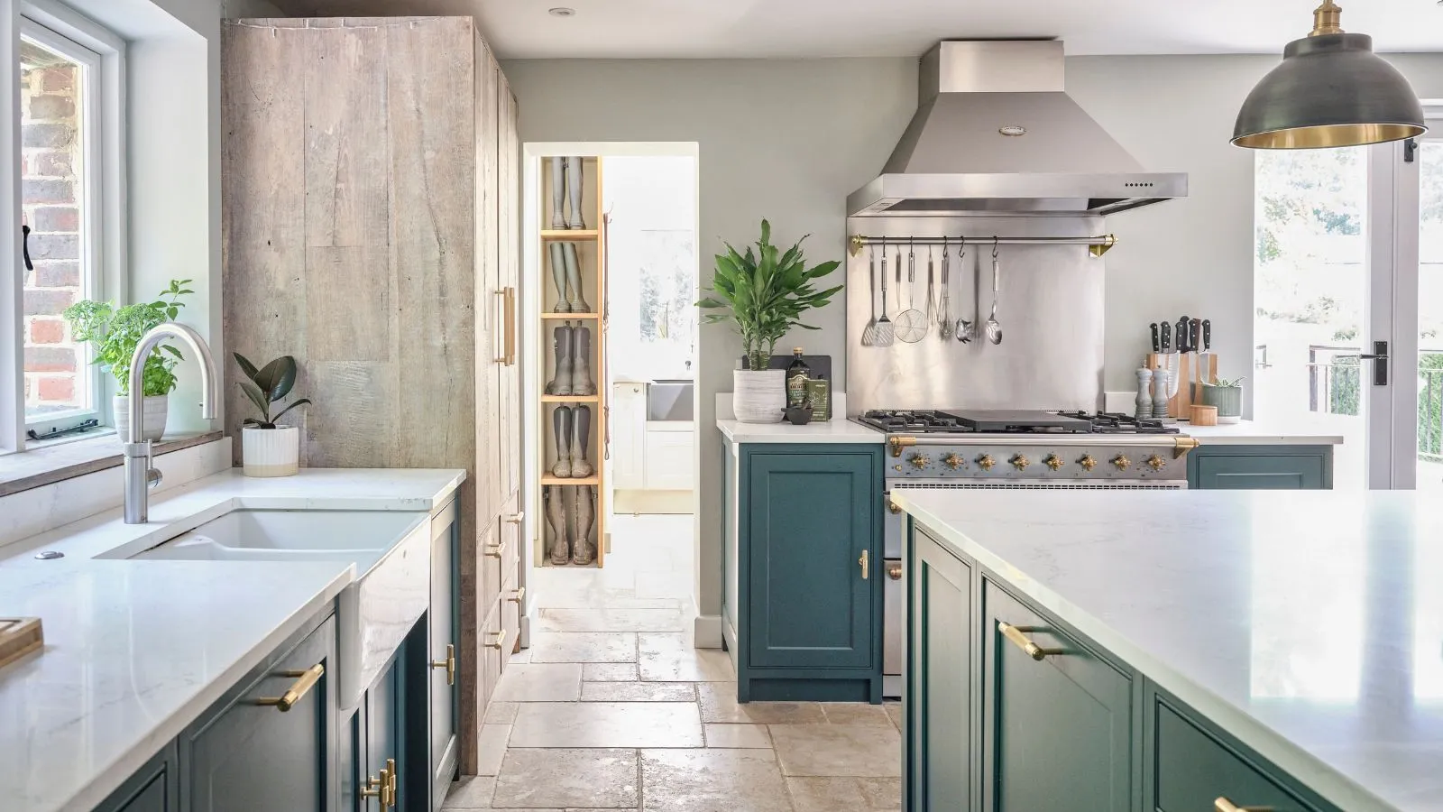 Uma cozinha leve e arejada com luzes pendentes de unidades azuis e piso de laje. Um backsplash de aço inoxidável e um ventilador de extrator atrás/acima do fogão.' loading='lazy' title=