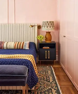 colori che si abbinano al rosa chiaro, camera da letto con pareti rosa chiaro, letto imbottito a righe, coperta blu indaco, comodino blu scuro, pouf blu scuro, tappeto vintage, pavimento in legno, lampada da parete, paralume fantasia sulla lampada da tavolo, cuscino da letto a righe, coperta a quadri curcuma, armadi