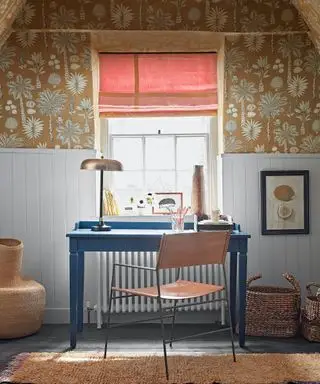 Escritorio azul con silla de cuero y metal, ventana con persiana roja, papel tapiz floral ocre, paneles pintados de azul claro, lámpara de mesa metálica, alfombra texturizada y cestas tejidas