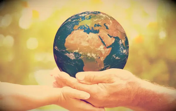 The Earth being held by one young hand and one old hand with a backdrop of green nature.