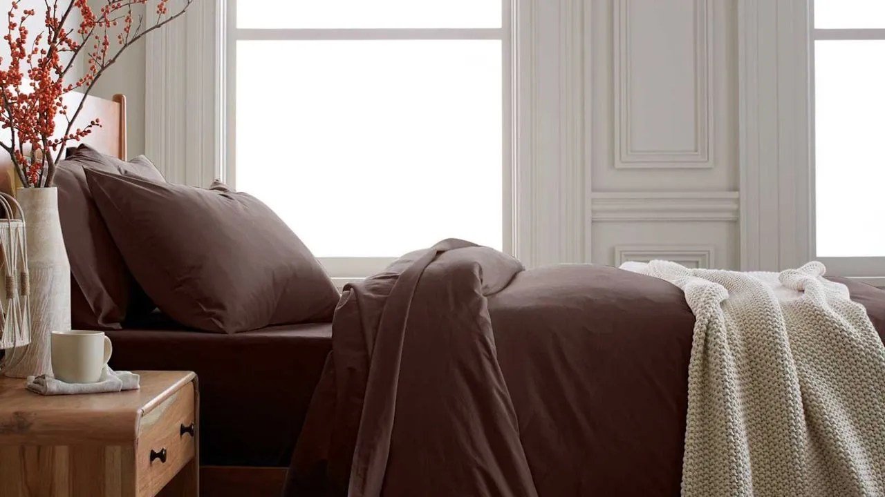 Mørkebrunt sengetøj på en træseng med et træ sengebord dekoreret med en vase med orange bær' loading='lazy' title=