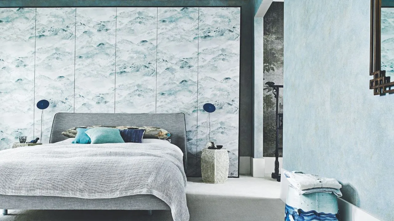 Une chambre bleue avec un lit double gris, un mur de papier peint, deux lampes bleues assorties et un tabouret à motifs bleus.' loading='lazy' title=