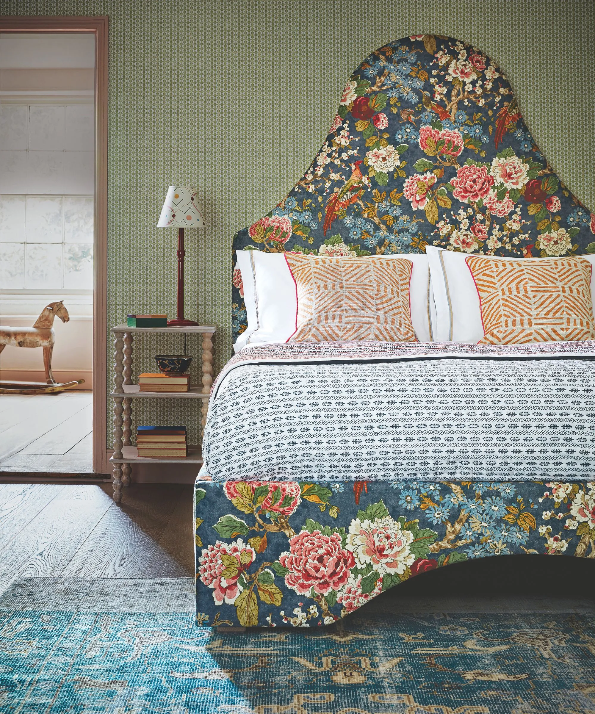 Une chambre vert maximaliste avec un grand lit à motifs floraux avec une tête de lit tendance et des coussins à motifs orange. À côté du lit se trouve une table de chevet en bois avec des livres et une lampe en bois surmontée d'un abat-jour blanc. Au premier plan se trouve un tapis à motifs bleus. Au fond, à gauche, une porte ouverte avec un cheval à bascule en bois visible au-delà.' loading='lazy' title=