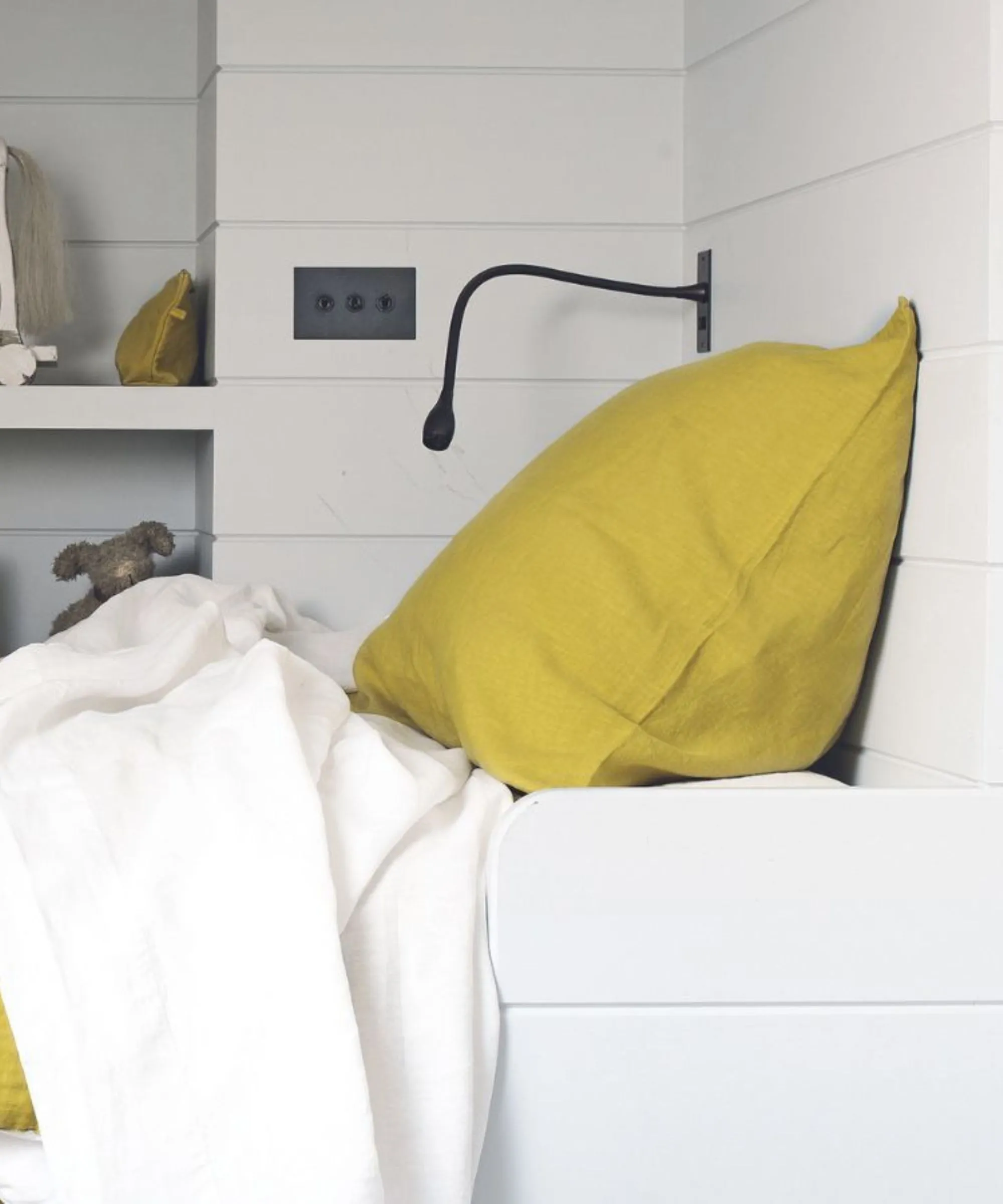 Une chambre lambrissée blanche avec un lit blanc avec un oreiller jaune, une lampe de lecture noire, des interrupteurs noirs et des étagères intégrées derrière avec un ours en peluche' loading='lazy' title=