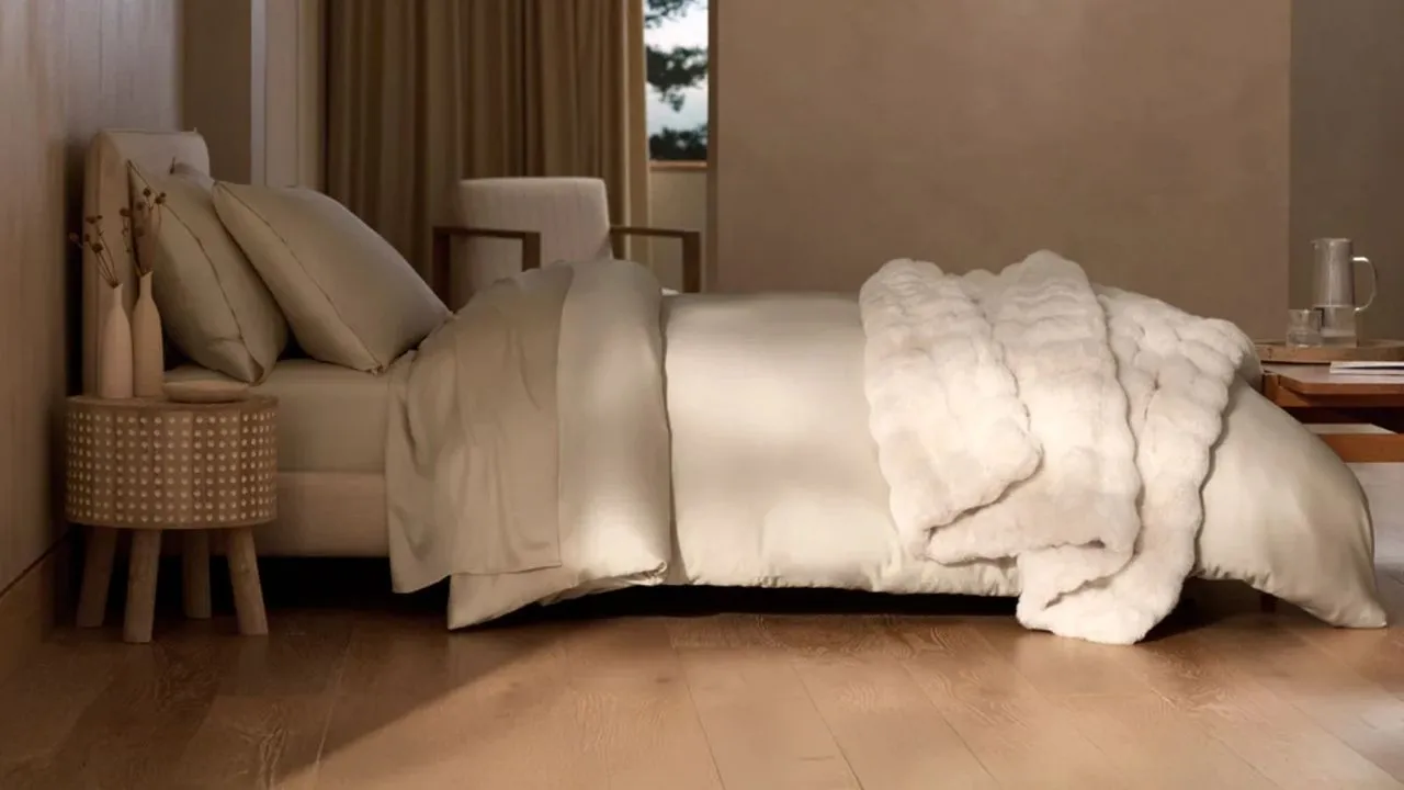 크림색 침구와 대형 더블 침대를 갖춘 현대적이고 미니멀한 침실입니다. 바닥은 나무로 되어 있고 전경의 침대 오른쪽에는 말린 꽃이 담긴 꽃병 두 개가 놓인 원형 침대 옆 탁자가 있습니다. 배경에는 갈색 커튼이 그려진 커다란 창문이 있습니다.' loading='lazy' title=