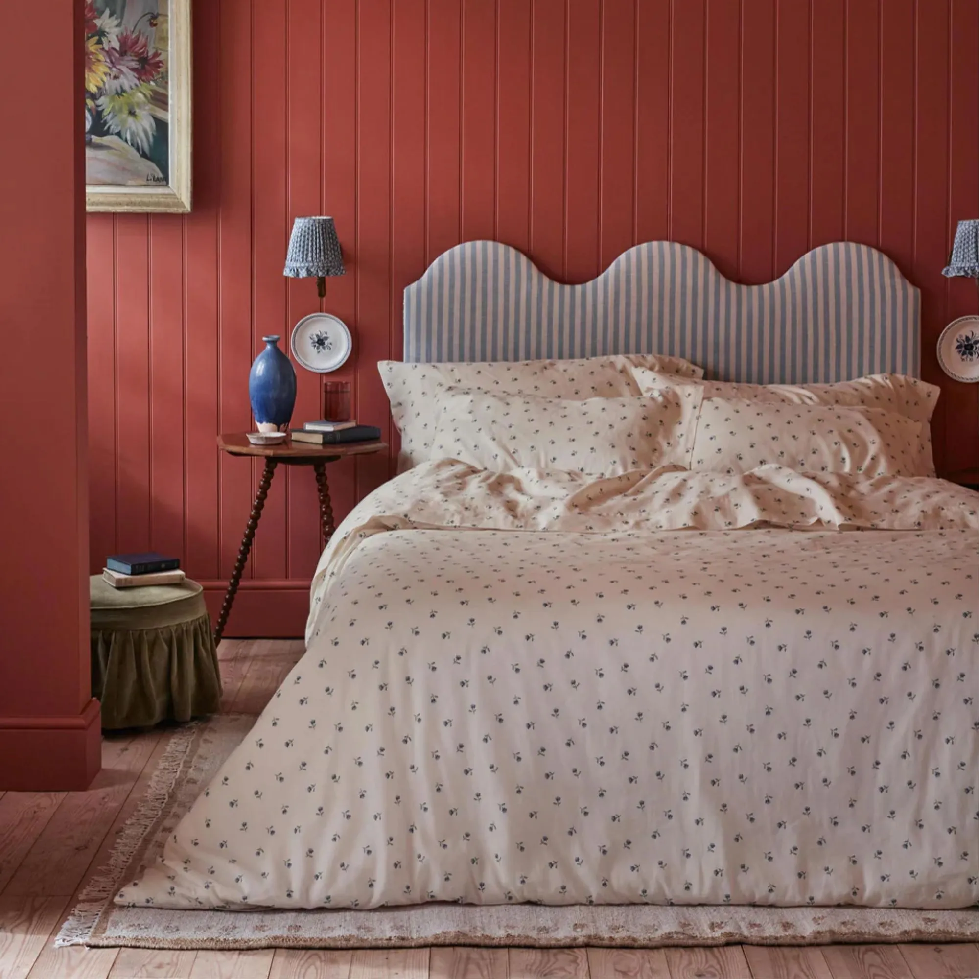 ベッドの中の子豚 ブルー ダスク ポージー 寝具、波型の青と白のストライプのヘッドボードを備えたダブルベッド。部屋には深紅のパネルがあり、ベッドの横には青い花瓶が付いた木製の円形テーブルがあります。床は淡い色の木材で、ベッドの下には大きな敷物が敷かれています。' loading='lazy' title=