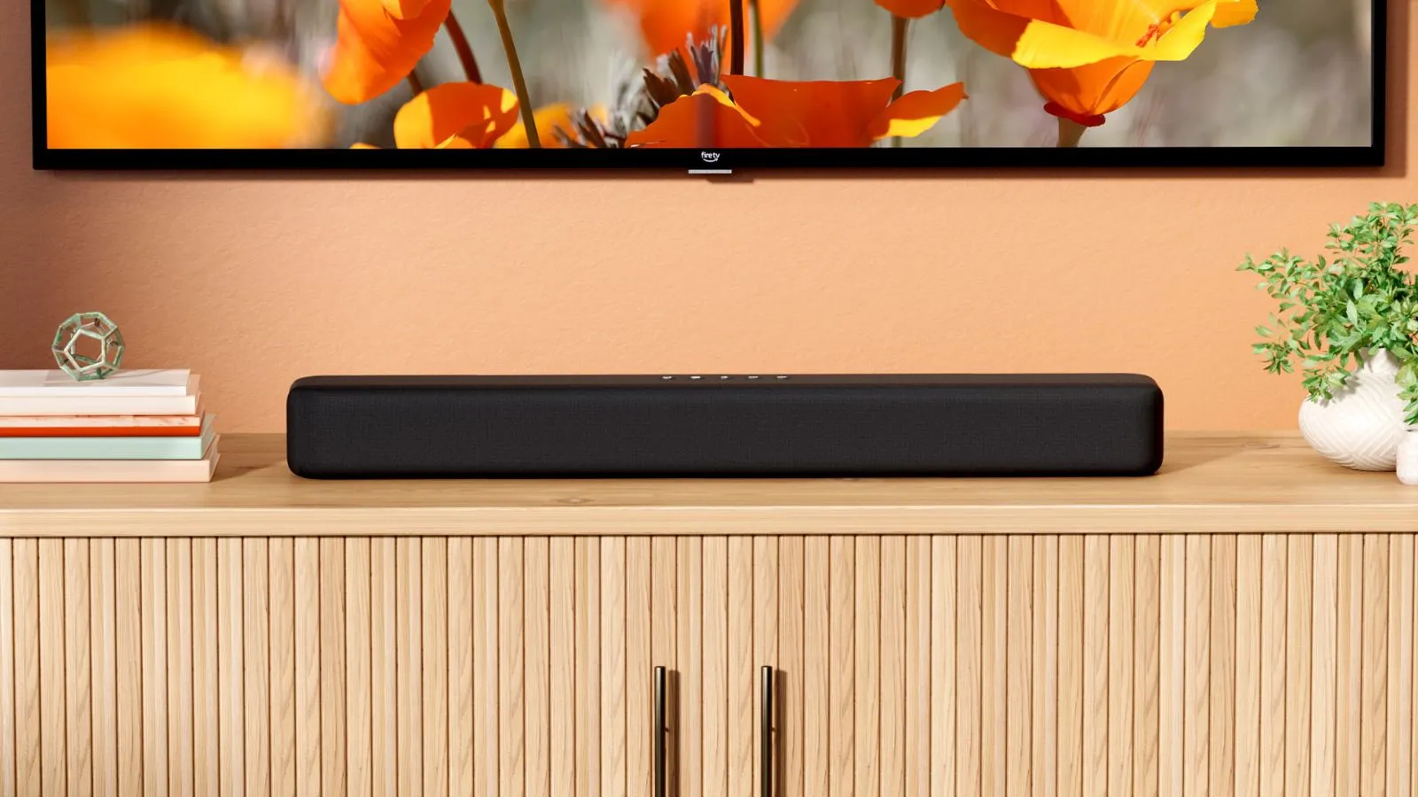 Soundbar Amazon Fire TV pod ekranem telewizora przedstawiający pomarańczowo-czerwone kwiaty na drewnianym stojaku pod telewizor obok książek i roślin na pomarańczowej ścianie.' loading='lazy' title=