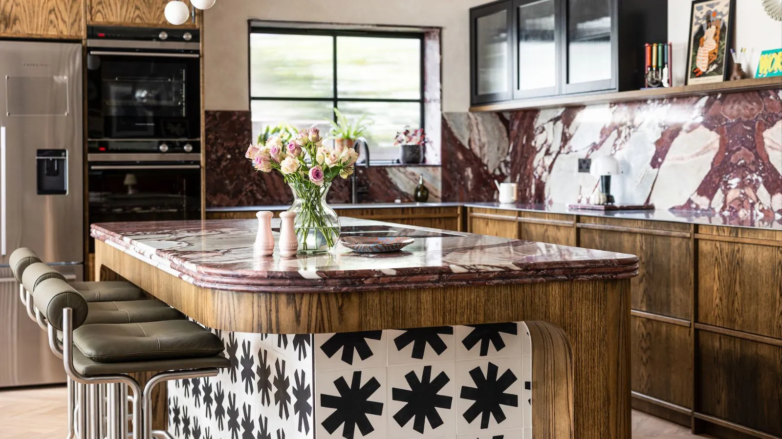 Una cucina con mobili rustici in legno e ripiani e alzatine in marmo rosso. Un'isola coordinata con rivestimento in piastrelle nere e bianche.' loading='lazy' title=