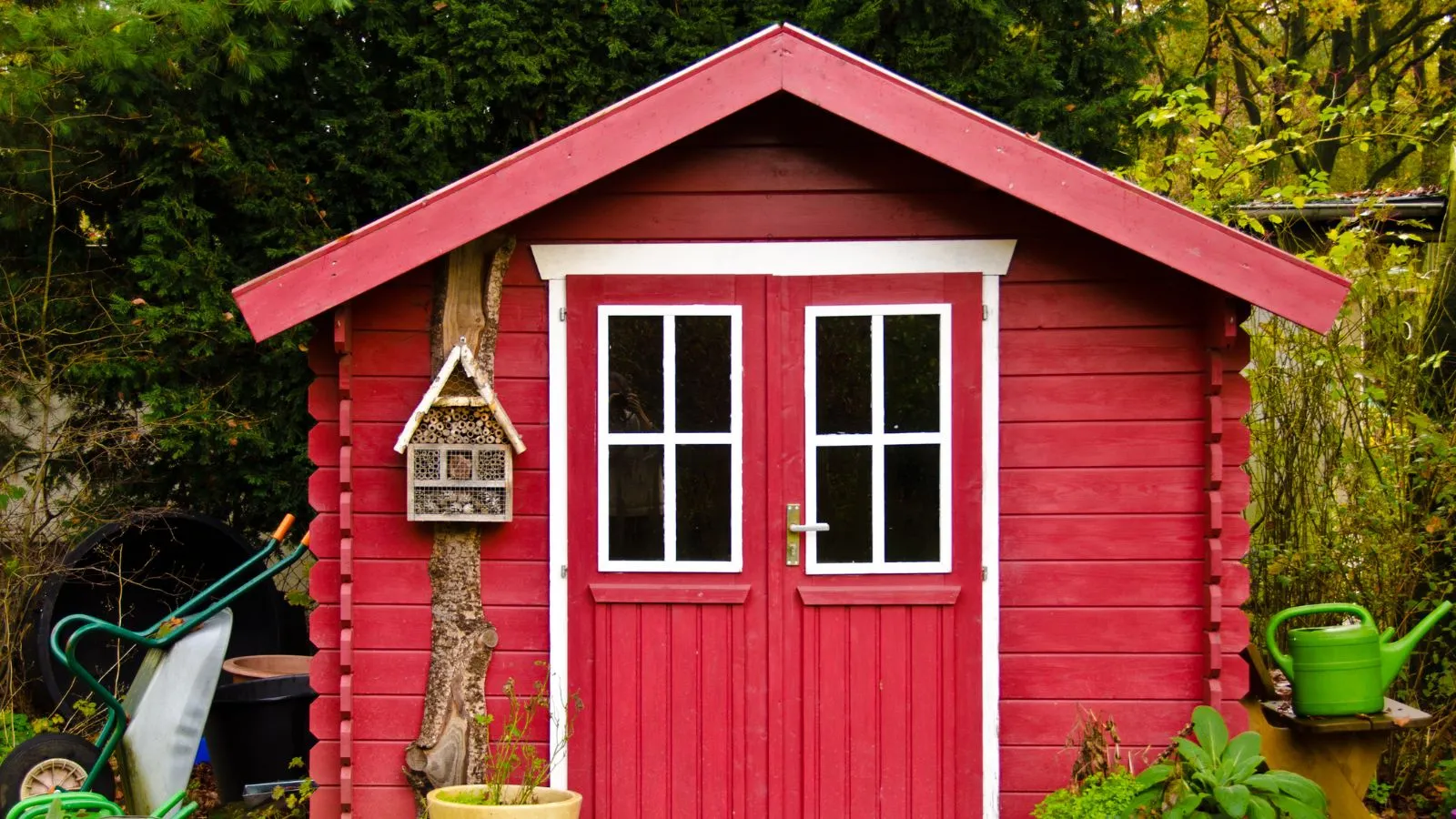 Una piccola casetta da giardino rossa circondata da cespugli verdi, alberi e attrezzi da giardino sparsi.' loading='lazy' title=