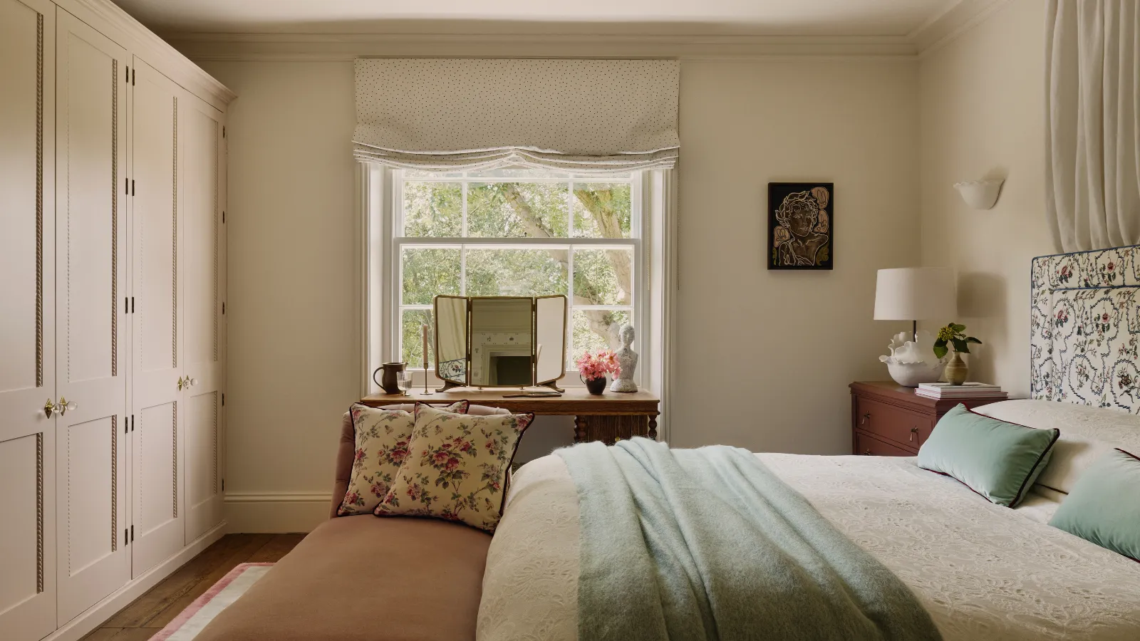 ein Schlafzimmer mit neutralen Wänden, einer Schrankwand, einem großen Fenster, einem Bett mit cremefarbener Bettwäsche, einer blaugrünen Decke und blaugrünen Kissen sowie einer Bank am Fußende des Bettes mit zwei geblümten Kissen' loading='lazy' title=