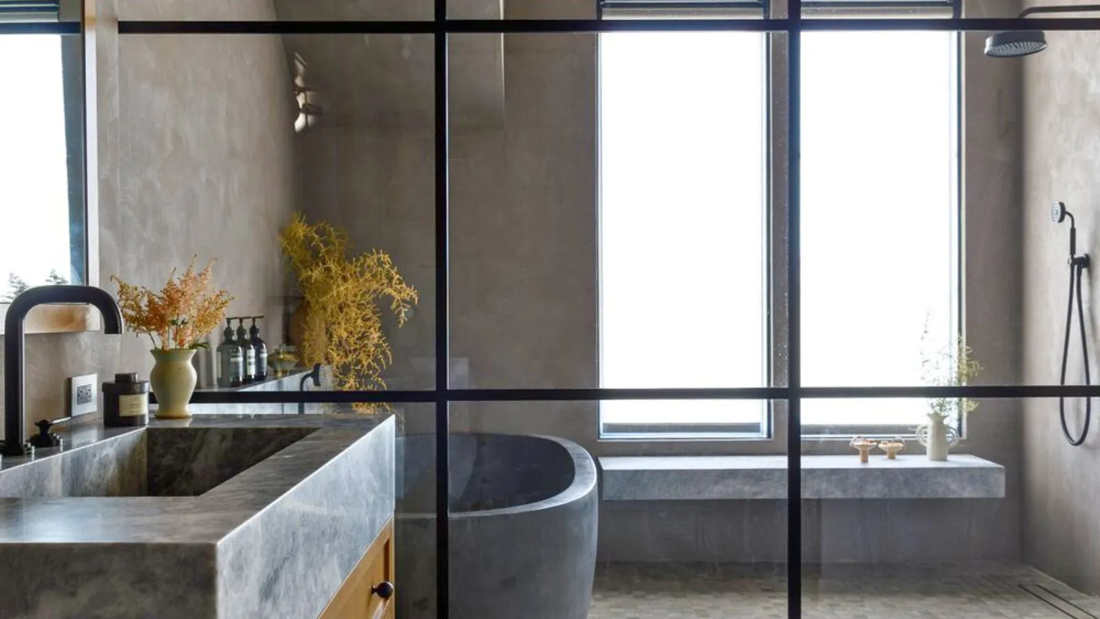 Szklany prysznic Crittal w łazience z neutralnego kamienia. Widoczne jest siedzisko prysznicowe i duże oszronione okno, a także wanna z szarego kamienia i kamienna umywalka' loading='lazy' title=