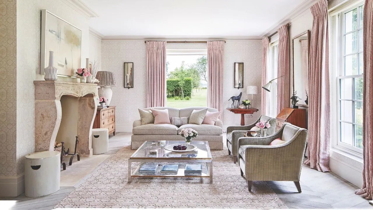 En rosa stue med rosa gardiner et rosa mønstret teppe i glass salongbord nøytrale sofaer og en stor steinpeis. Veggene er hvite og en grønn hage kan sees gjennom vinduene langs veggene.' loading='lazy' title=