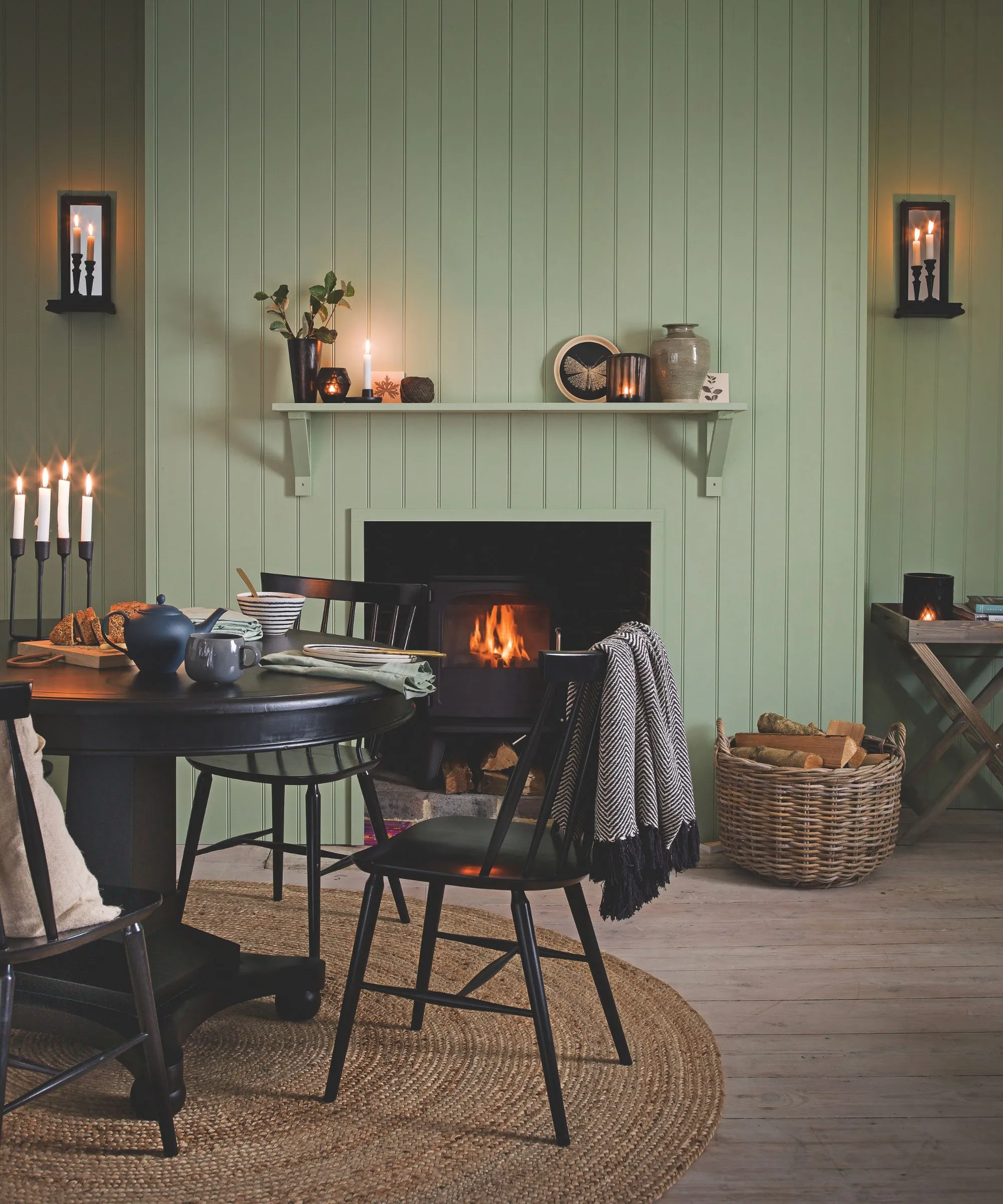 En grønn panelt stue med tent bål i peisen rotting sirkulært teppe sirkulært svart spisebord med matchende stoler en flettet kurv av ved og tente koniske lys.' loading='lazy' title=