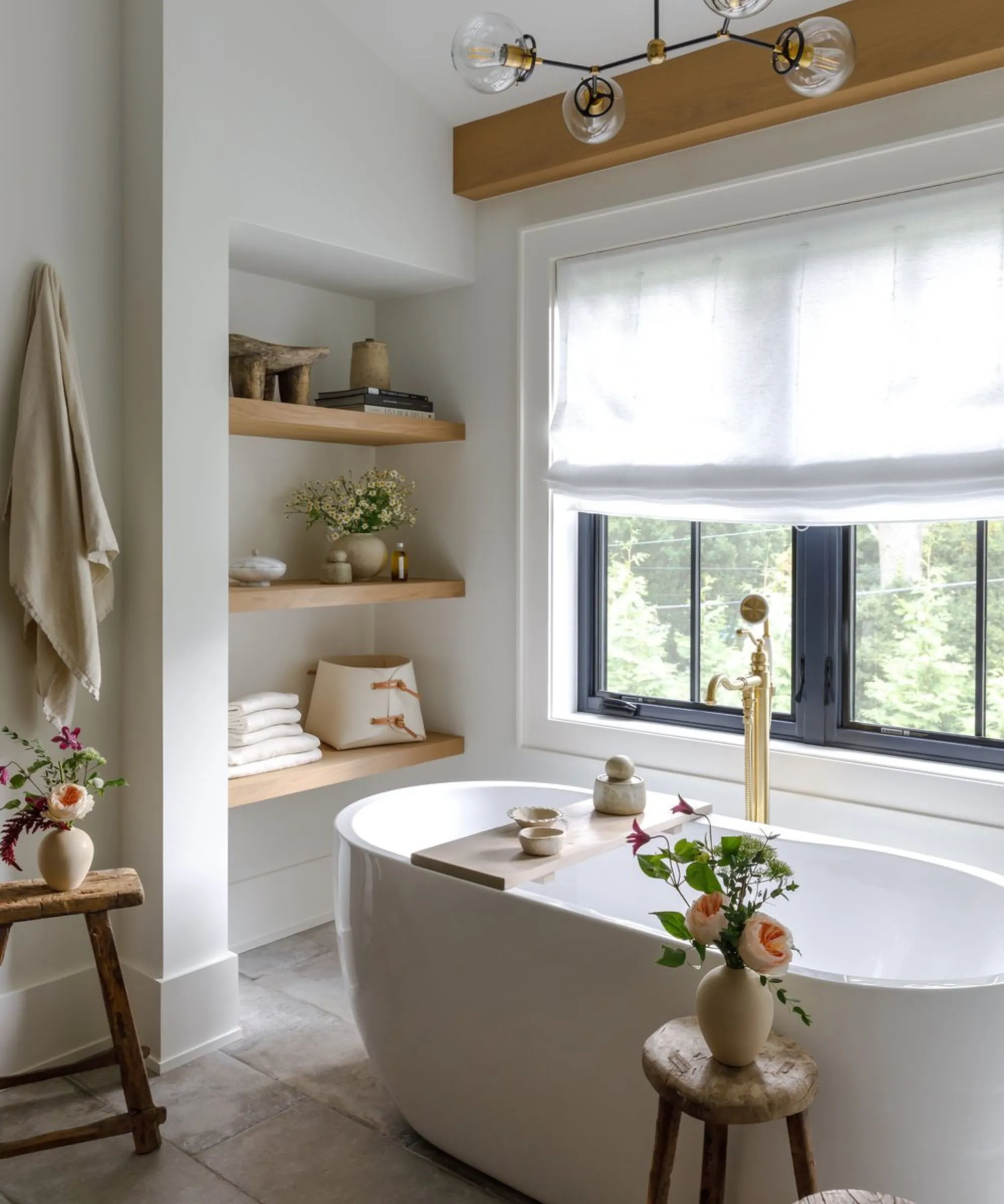 Mała biała, nieskazitelnie czysta łazienka z wolnostojącą wanną i półkami. Znajdują się tam drewniane stołki z wazonami z kwiatami, a także bulwiasta lampa sufitowa i lniane dekoracje okienne' loading='lazy' title=