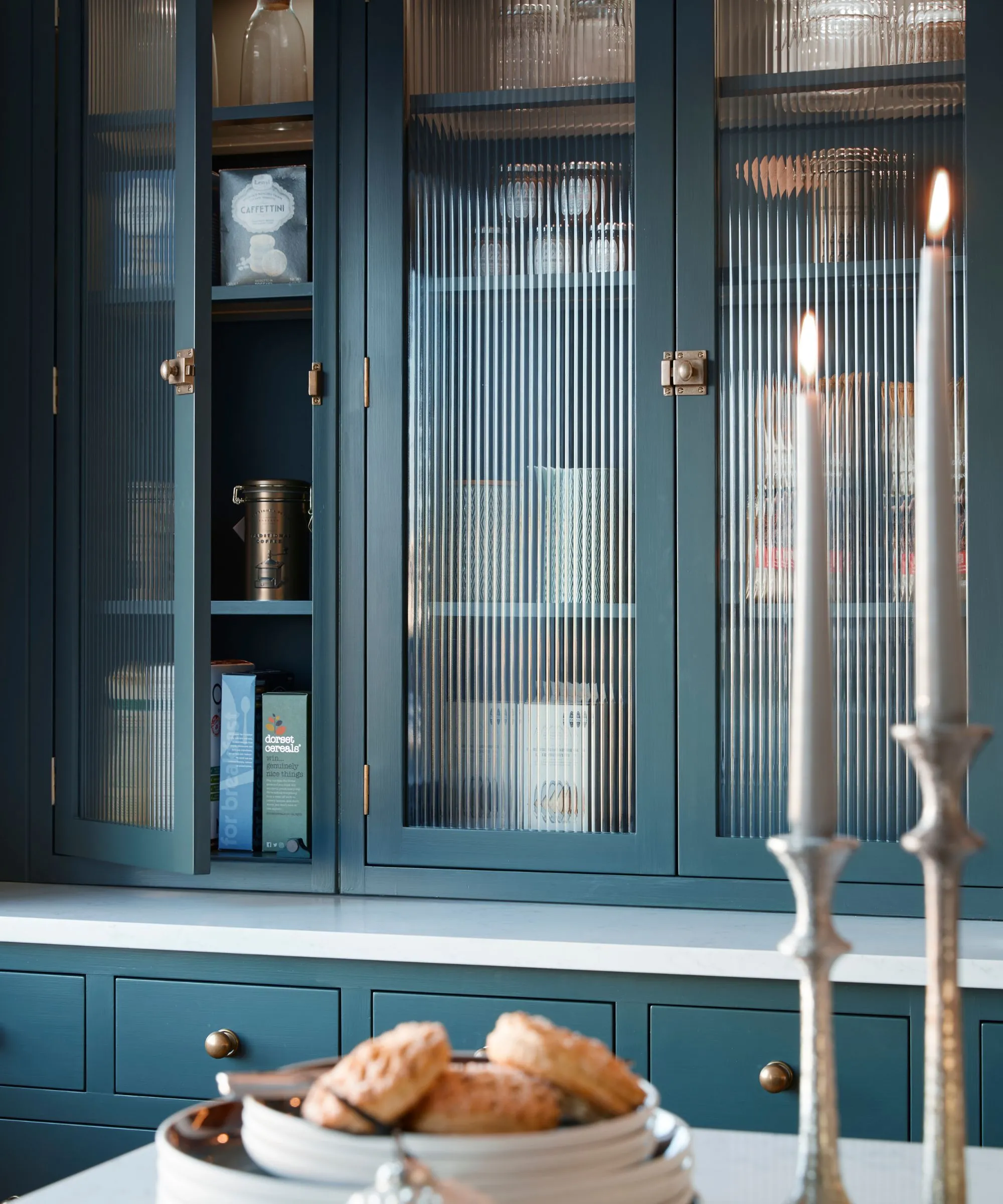 Duże niebieskie szafki kuchenne ze szklanymi frontami. Jedne drzwi częściowo otwarte, aby pokazać składniki spiżarni. Na pierwszym planie rozmazany talerz małych ciastek i dwie zapalone świece.' loading='lazy' title=