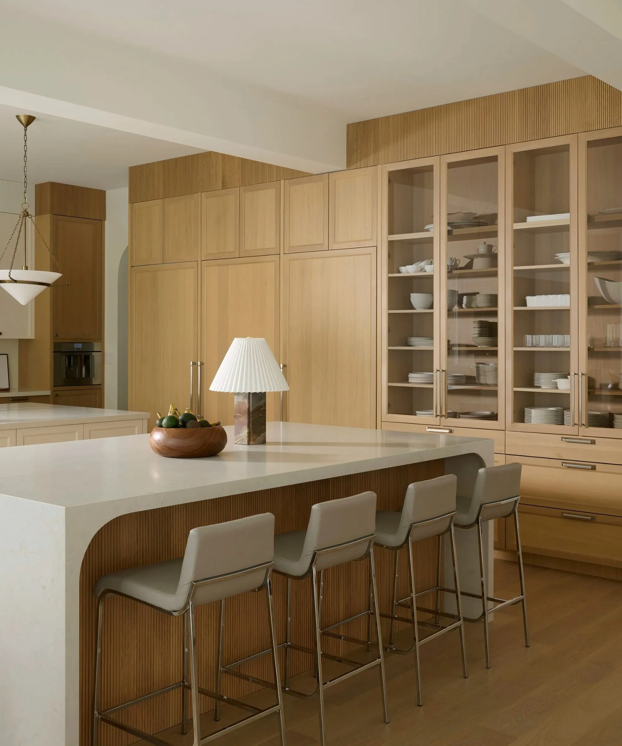 uma cozinha tranquila com uma parede de armários embutidos uma ilha de cozinha com bancada branca quatro banquetas e uma luminária decorativa e fruteira na ilha' loading='lazy' title=