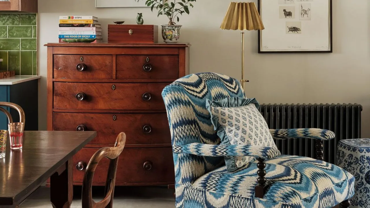 Et åpent kjøkken med spisestue med bare et hjørne av et kjøkkenskap i sikte med grønn T-bane-splashback fliser. Foran og i midten er en mønstret, polstret lenestol i blått og kremfarget med tretrekk. I bakgrunnen er det en svart radiator synlig samt et skap med fem skuffer fra midten av århundret' loading='lazy' title=