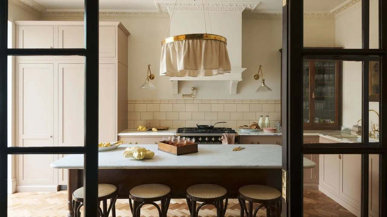 Karakter ferskenrosa kjøkken sett gjennom innvendige svarte Crittal-dører. Det er et marmorbord med ryggløse barkrakker av tre i sikte. I bakgrunnen er det en splashback kakkelovnsplass og innebygde rosa og glassfrontbrune skap.' loading='lazy' title=